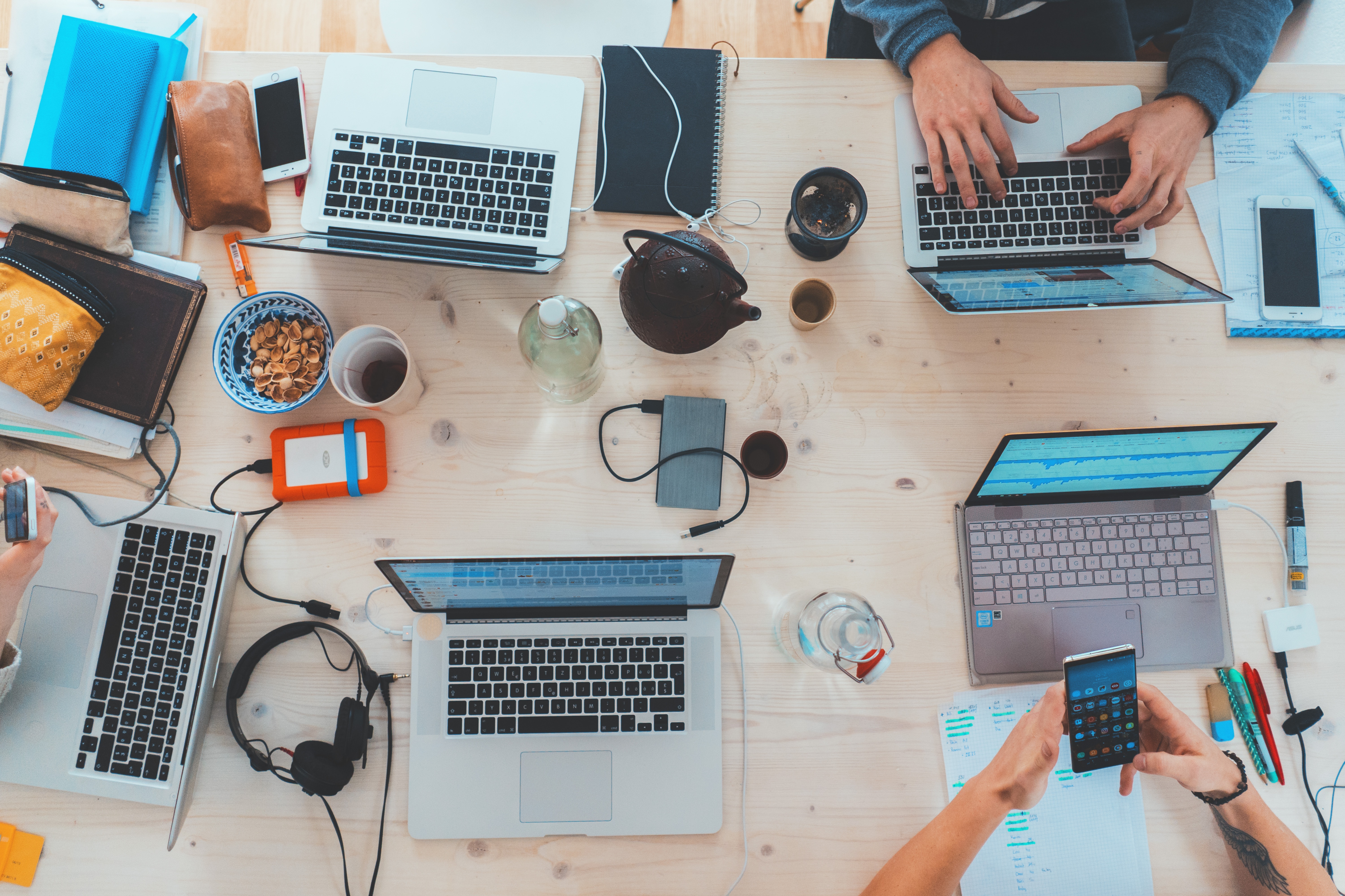 Pięć laptopów z otwartymi ekranami na stole. W różnych orientacjach (obok, przed, pomiędzy) lezą też inne przedmioty - słuchawki, smartfony, ładowarki, kubki.