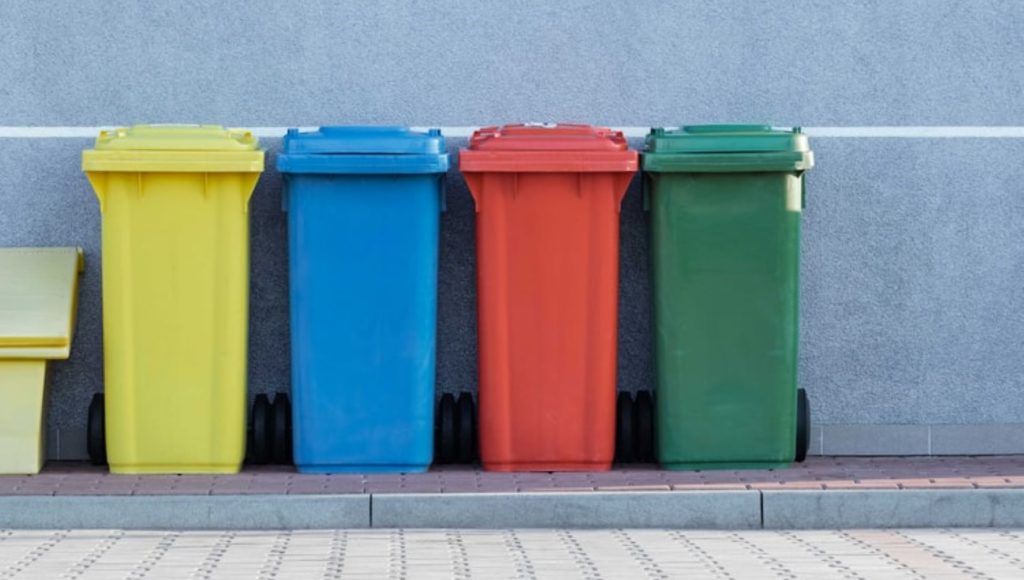 Cztery kosze na śmieci stojące przy ścianie. Od lewej: żółty, niebieski, czerwony i zielony.
