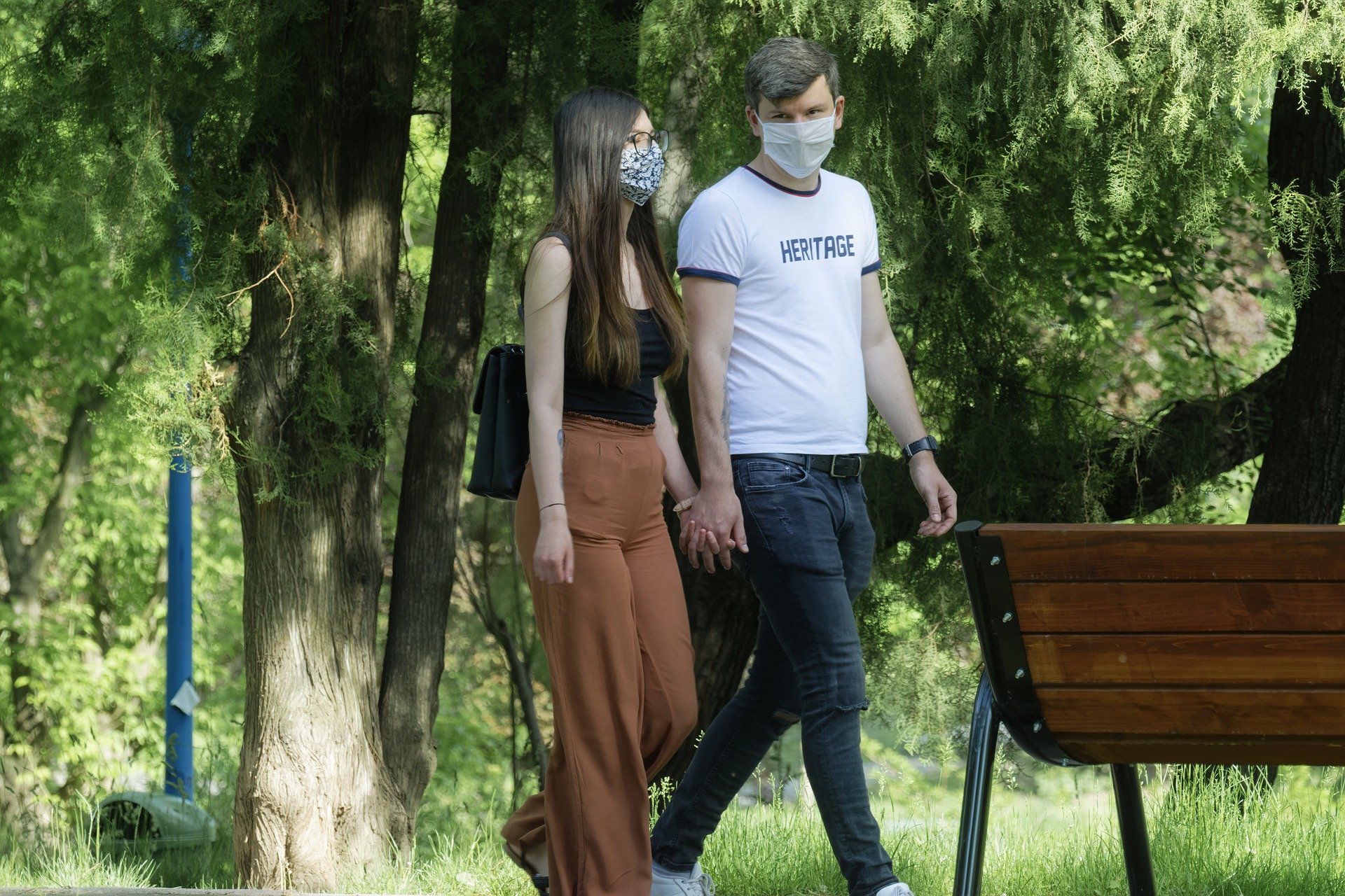 Mężczyzna i kobieta spacerują po parku, trzymając się za ręce. Oboje mają na twarzach maseczki ochronne.
