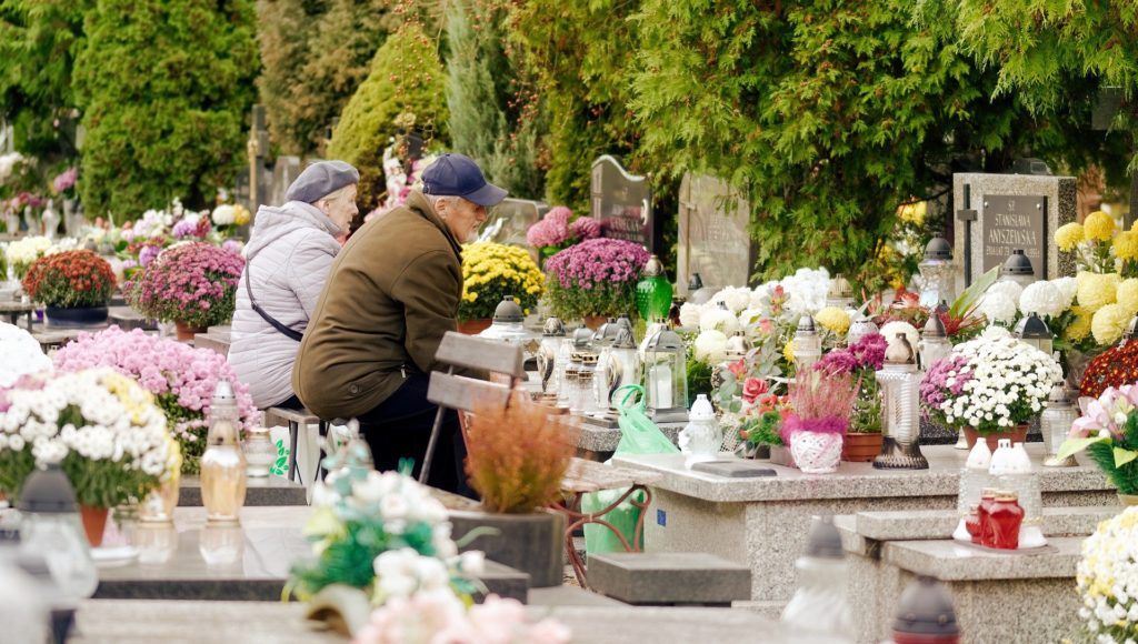 para starszych ludzi siedzi na ławce przed nagrobkiem na cmentarzu, na grobie jest pełno kwiatów i zniczy