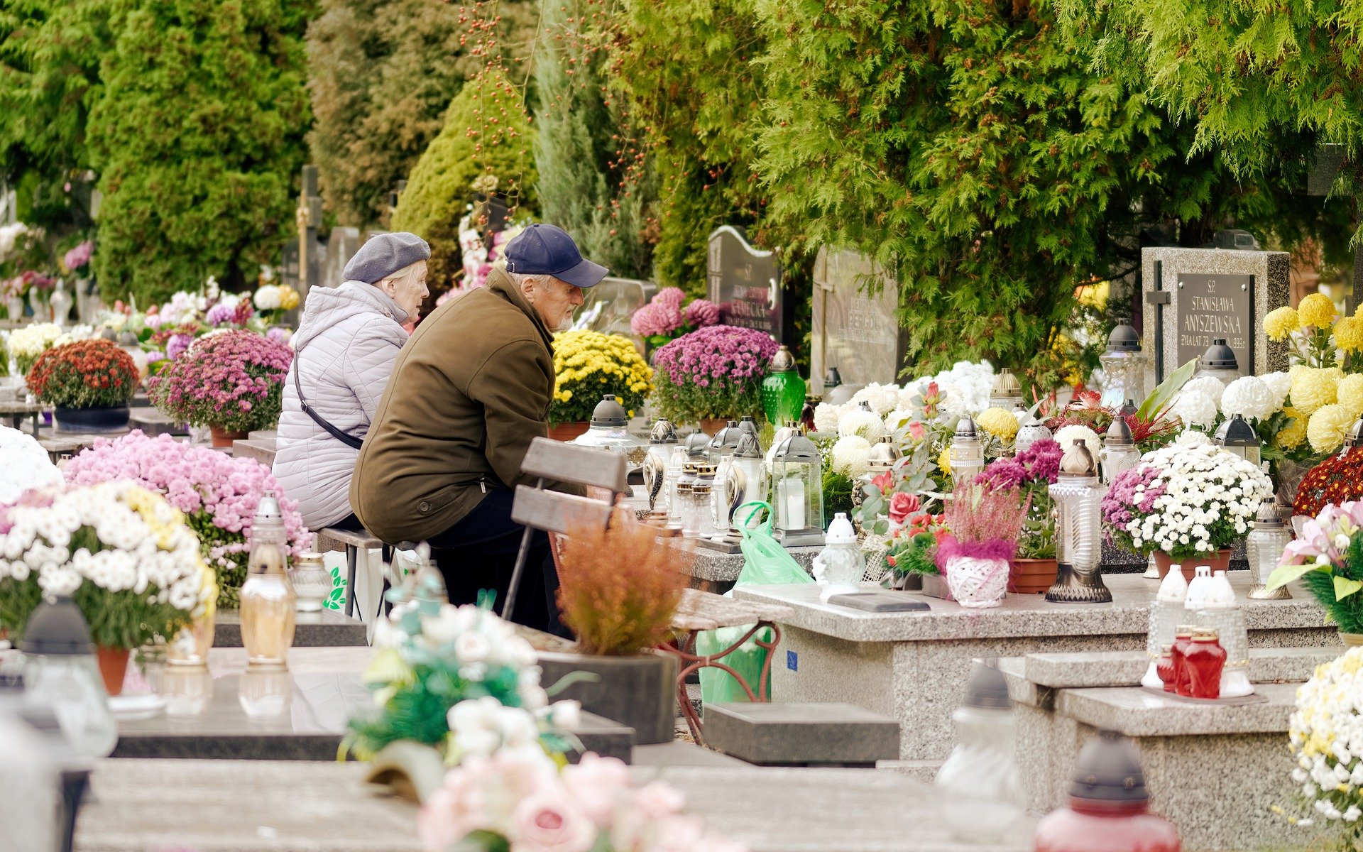 para starszych ludzi siedzi na ławce przed nagrobkiem na cmentarzu, na grobie jest pełno kwiatów i zniczy