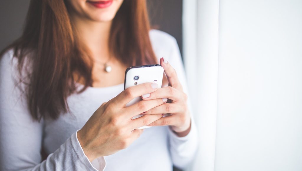 kobieta trzyma telefon zwrócony w swoją stronę, widać tylko dolną połowę jej twarzy oraz korpus