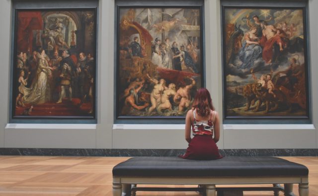 młoda kobieta siedzi na ławce w muzeum i patrzy na wiszące przed nią 3 obrazy; widzimy ją od tyłu