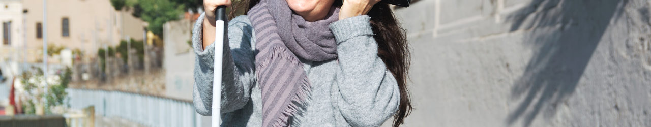 Uśmiechnięta kobieta siedzi na murku. W jednej ręce trzyma białą laskę, w drugiej, tuż przy uchu, smartfon.