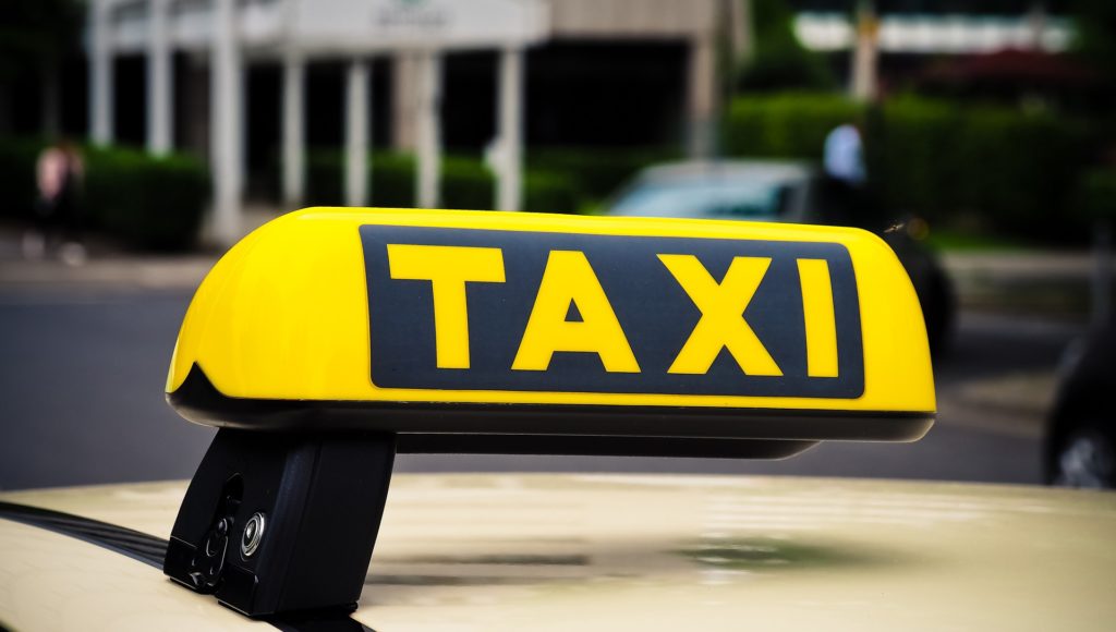 Do dachu samochodu przyczepiony żółto-czarny napis TAXI (żółte litery na czarnym tle).