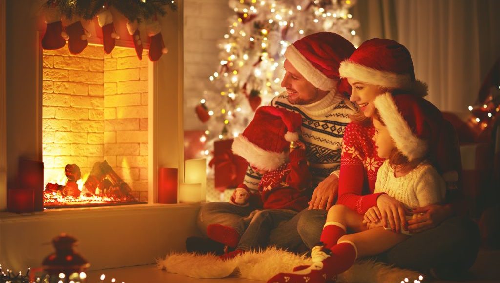 rodzina w czapkach Mikołaja siedzi razem przed kominkiem, rodzice trzymają dzieci na kolanach, wszyscy uśmiechają się, w tle choinka