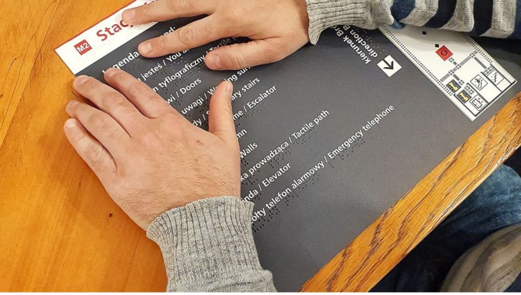 Dłoń osoby niewidomej dotykająca napisu brajlowskiego na tablicy informacyjnej