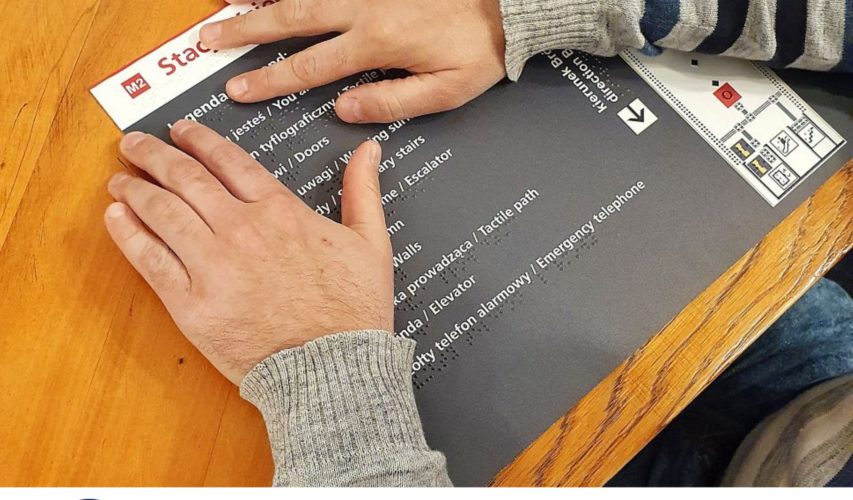 Dłoń osoby niewidomej dotykająca napisu brajlowskiego na tablicy informacyjnej