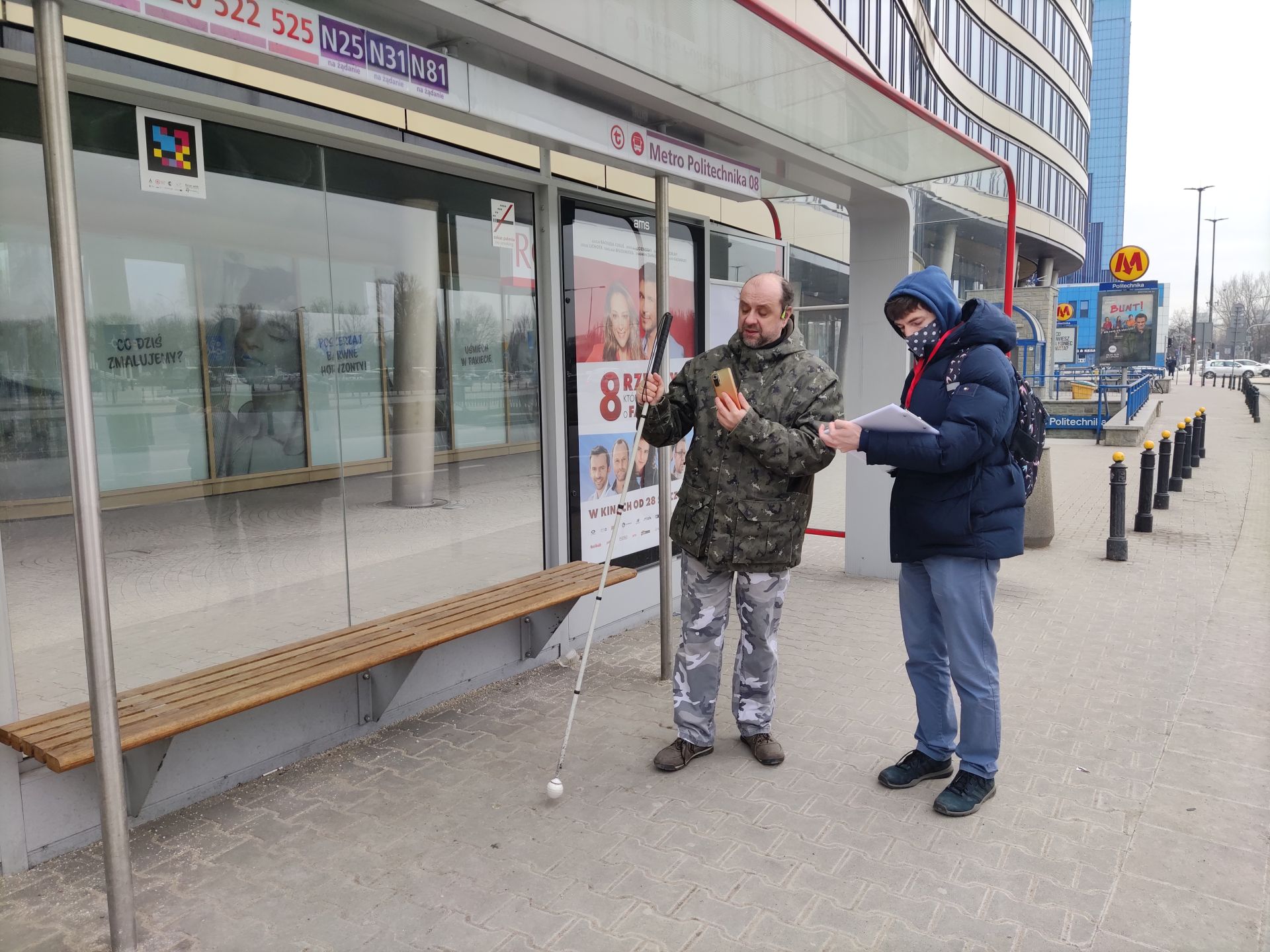 tester stoi na przystanku Metro Politechnika, sprawdza aplikację, za nim stoi koordynator i robi notatki