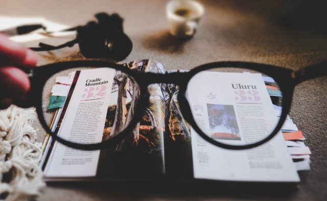 Zdjęcie okularów, przez które widać otwarte czasopismo. Na dalszym planie kubek z napojem.