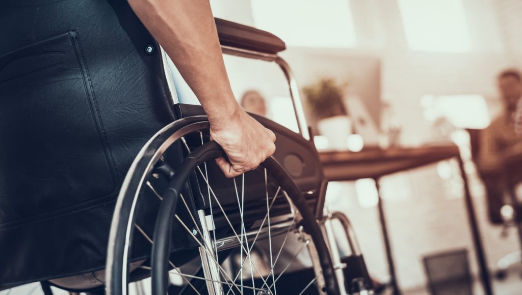 Osoba siedząca na wózku inwalidzkim, odwrócona tyłem, widać jej tylko prawą rękę. Dalsze tło rozmyte, widać na nim dwie osoby.