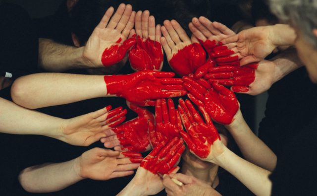 na zdjęciu widać ręce, które są ułożone blisko siebie i są pomalowane czerwoną farbą w taki sposób, że tworzą serce