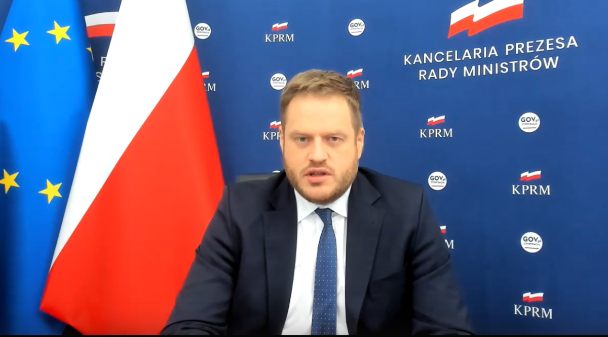 Janusz Cieszyński siedzi, za nim po lewej stronie flagi Polski i Unii Europejskiej