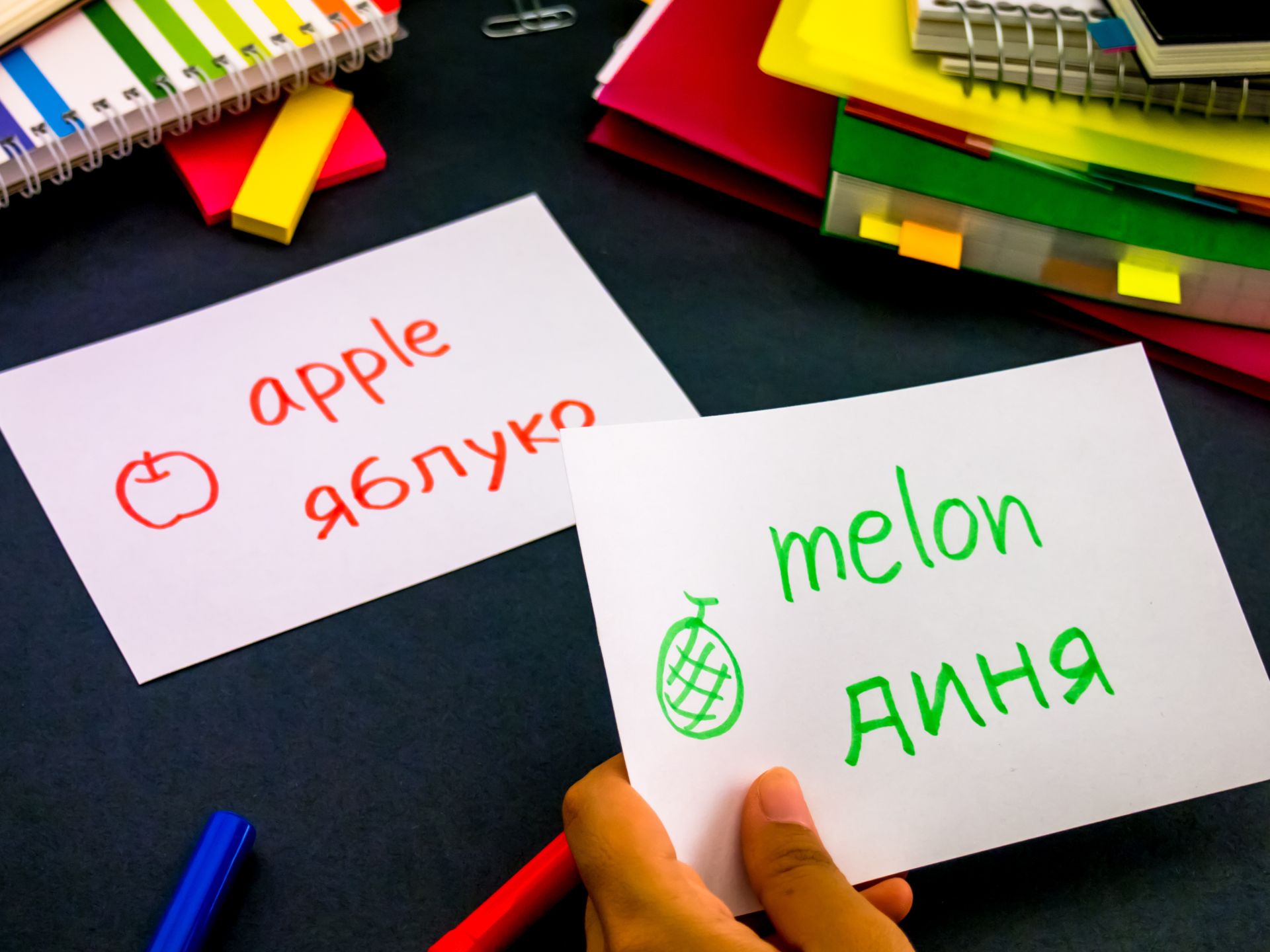 dużo książek i notatek na stole, zbliżenie na rękę, która trzyma kartkę ze słowem "melon" zapisanym po angielsku i ukraińsku, w tle taka sama karteczka, ale ze słowem "jabłko"
