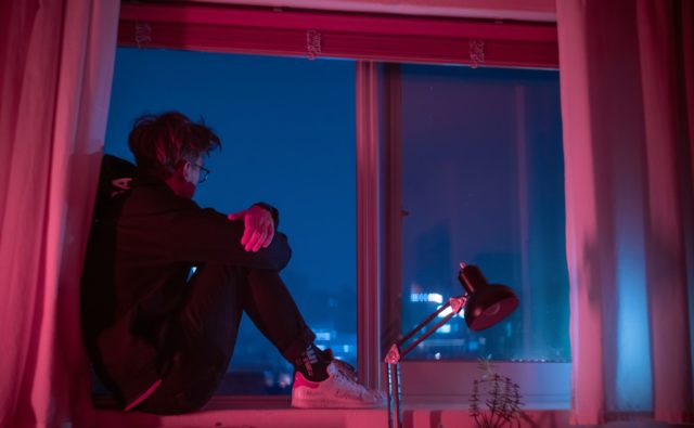 Młody mężczyzna siedzący nocą na parapecie i patrzący przez okno.