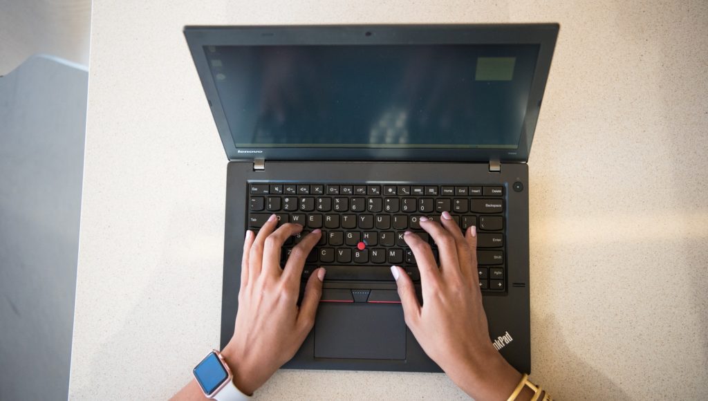 Zdjęcie zrobione z góry. Na biurku znajduje się laptop i dłonie kobiety piszące na klawiaturze.