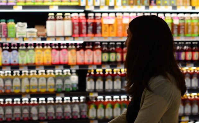 kobieta robi zakupy, stoi przed półką z napojami, które są lekko podświetlone, kobieta stoi trochę w cieniu, bokiem, widać ją z profilu
