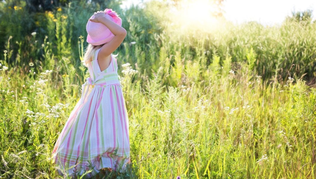 dziewczynka w długiej sukience i kapeluszu stoi na środku łąki, patrzy w stronę słońca