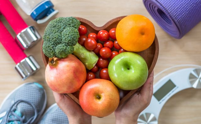 miska w kształcie serca w warzywami i owocami w środku, naokoło niej: waga, mata do ćwiczeń, hantle, ręcznik