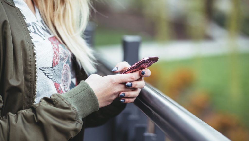 kobieta trzyma w rękach telefon komórkowy, nie widać jej twarzy, zbliżenie przede wszystkim na ręce; stoi w parku