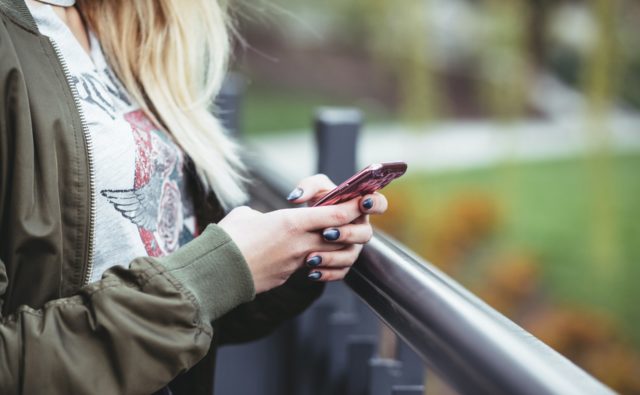 kobieta trzyma w rękach telefon komórkowy, nie widać jej twarzy, zbliżenie przede wszystkim na ręce; stoi w parku