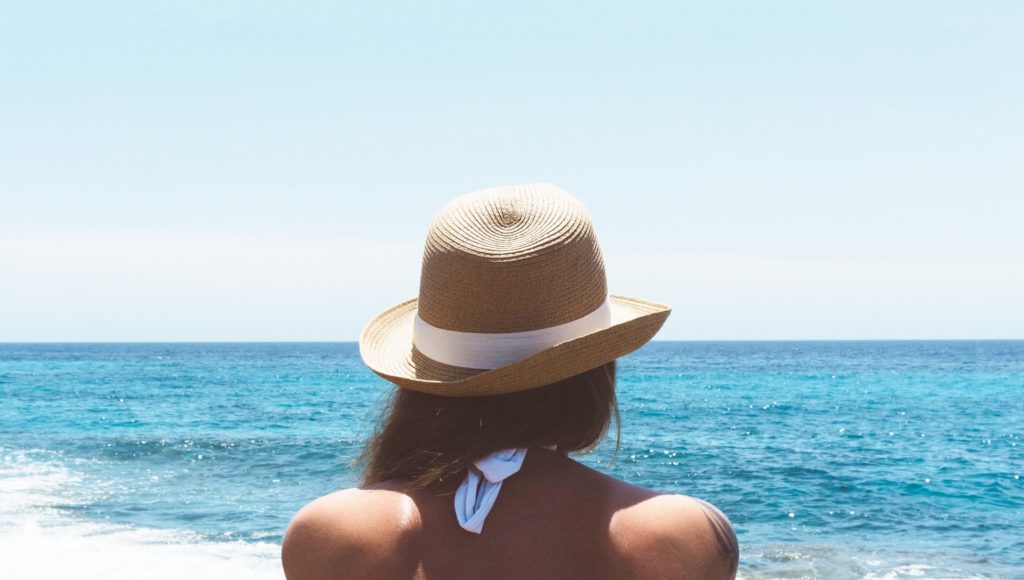 Kobieta w letnim kapeluszu i kostiumie kąpielowym siedzi i patrzy na morze; widzimy ją od tyłu, tylko do połowy pleców; jest słoneczna pogoda