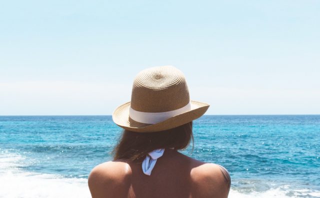 Kobieta w letnim kapeluszu i kostiumie kąpielowym siedzi i patrzy na morze; widzimy ją od tyłu, tylko do połowy pleców; jest słoneczna pogoda