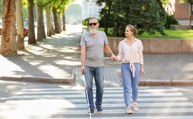 Starszy mężczyzna z białą laską przechodzi przez jezdnię. Pomaga mu dziewczyna.