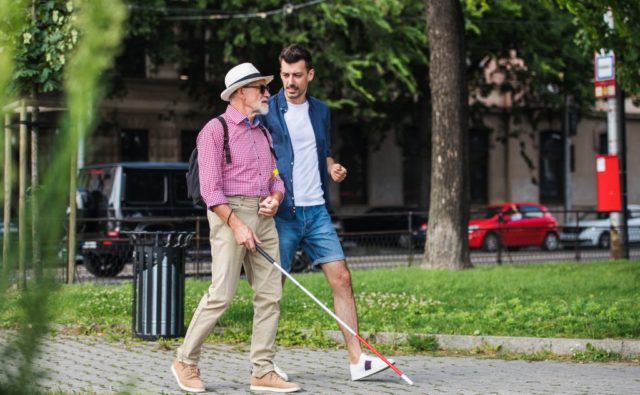 Ulicą idzie dwóch mężczyzn, jeden z nich ma białą laskę w dłoni.