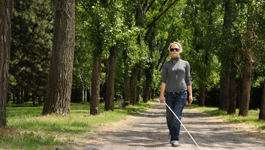 Niewidoma kobieta z białą laską idzie trasą pośród drzew