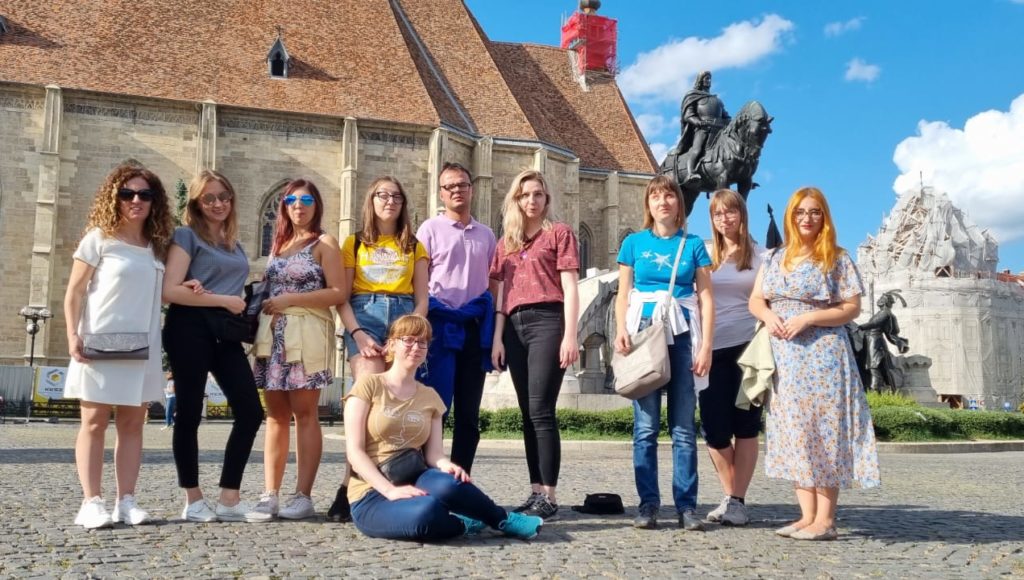 uczestnicy projektu, grupa młodych osób, stoi na placu przed kościołem w Rumunii i pozuje do zdjęcia; za nimi po prawej stronie jest pomnik przedstawiający postać na koniu