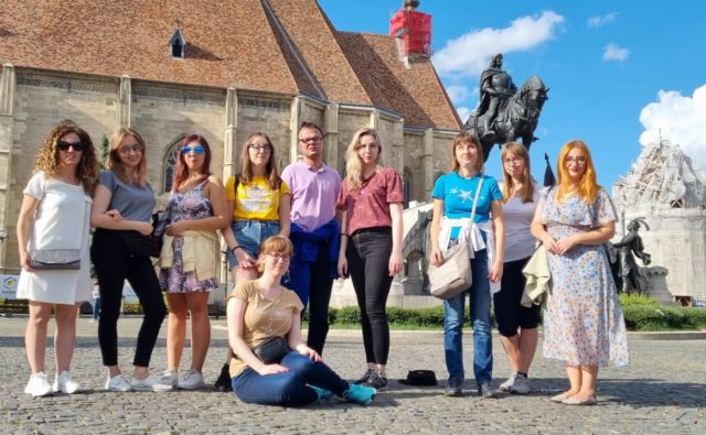 uczestnicy projektu, grupa młodych osób, stoi na placu przed kościołem w Rumunii i pozuje do zdjęcia; za nimi po prawej stronie jest pomnik przedstawiający postać na koniu