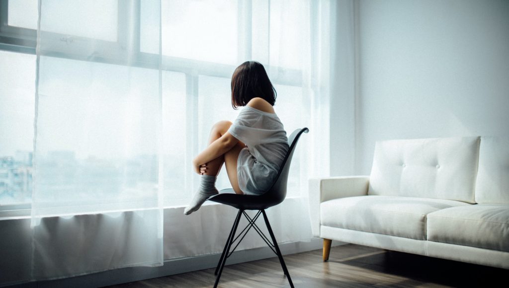 kobieta siedzi na krześle z podkulonymi nogami, patrzy przez okno, nie widać jej twarzy; za nią stoi biała kanapa