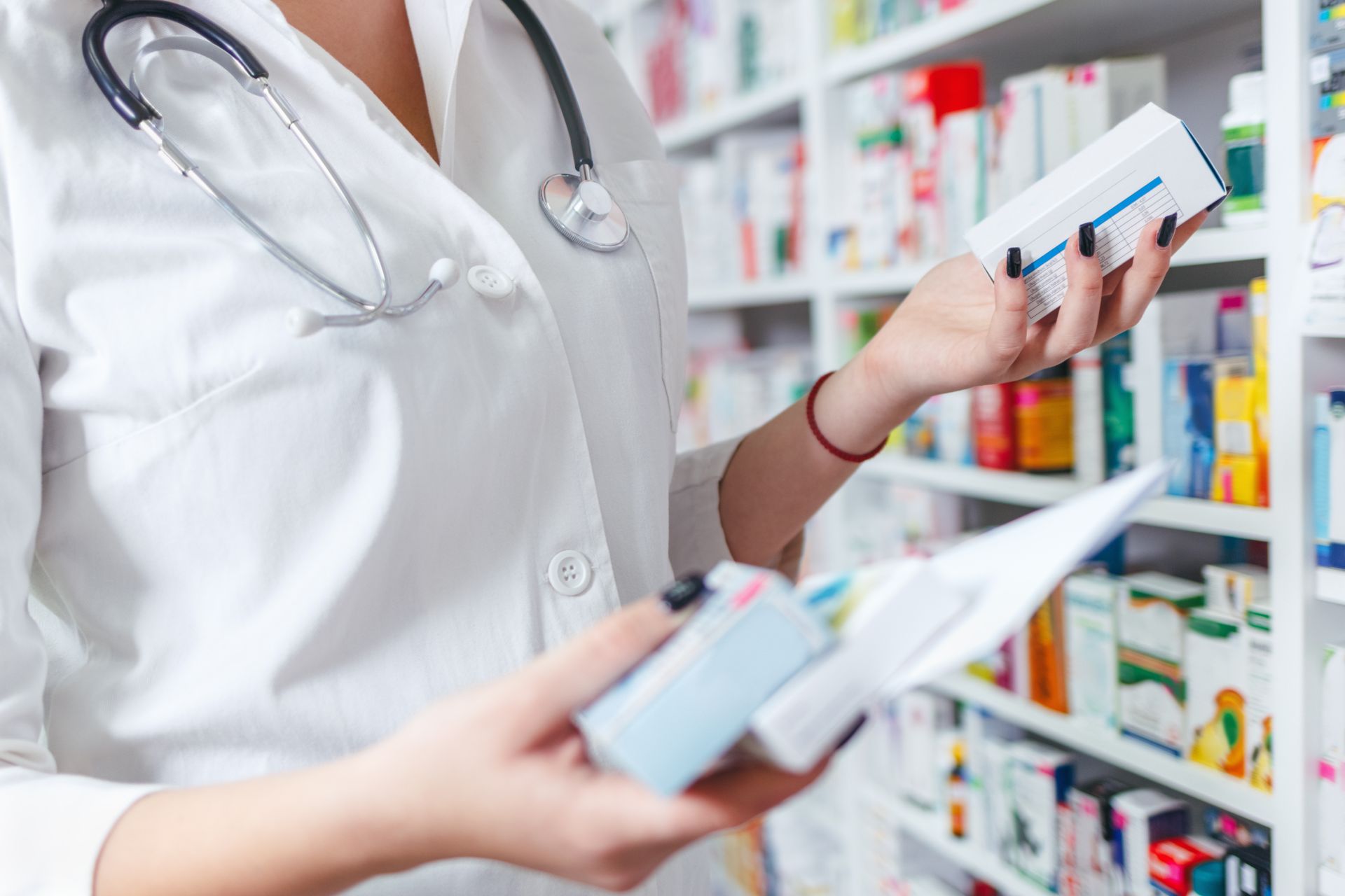 farmaceuta stoi przed półką z lekami i trzyma w dłoni opakowania z lekami, zbliżenie na ręce, nie widać jej twarzy