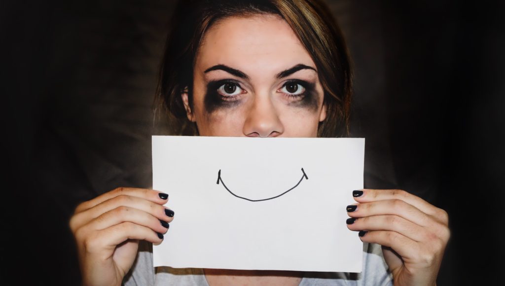 Na zdjęciu młoda kobieta z rozmazanym tuszem wokół oczu, trzyma kartkę z narysowanym uśmiechem na wysokości ust.