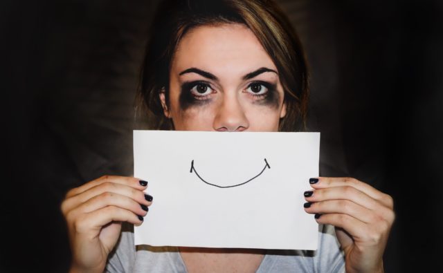 Na zdjęciu młoda kobieta z rozmazanym tuszem wokół oczu, trzyma kartkę z narysowanym uśmiechem na wysokości ust.