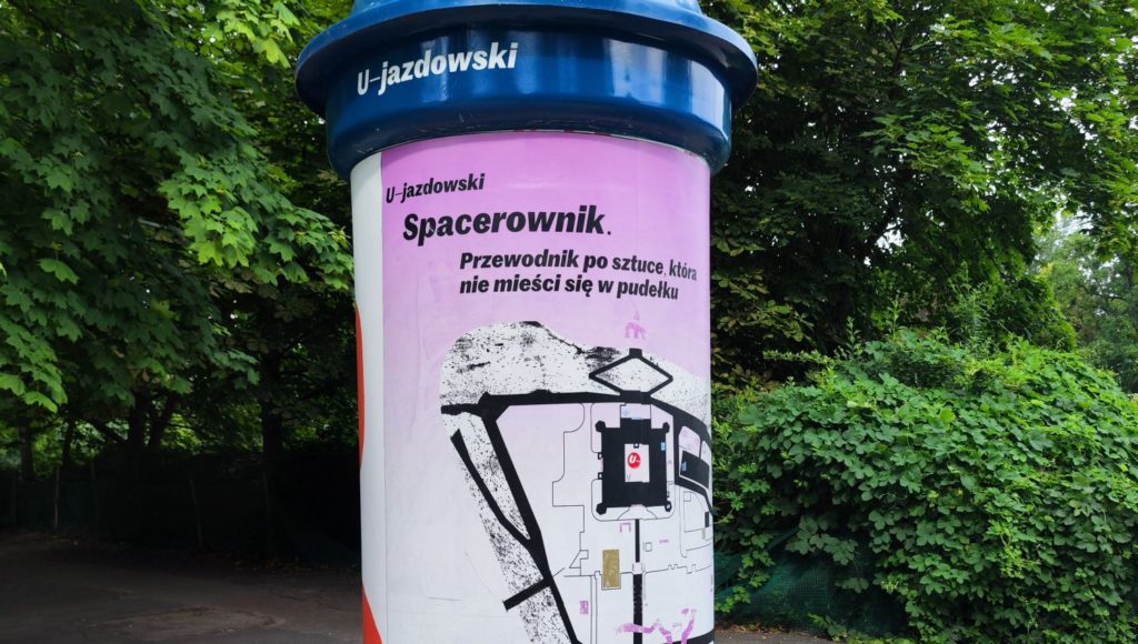 Słup informacyjny przy Parku Ujazdowskim z zawieszonym plakacie dotyczącym Spacerownika.
