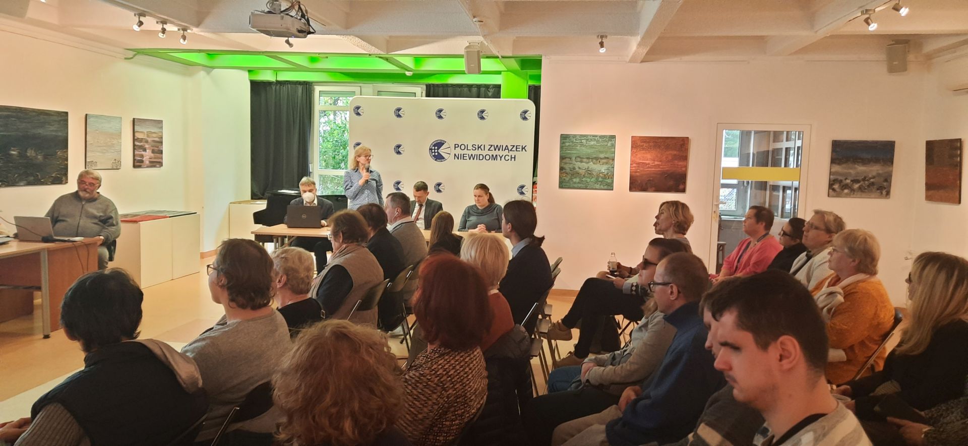 uczestnicy spotkania siedzą na krzesłach w Tyflogalerii, przy stole obok nich stoi Anna Lemańczyk i mówi do publiczności, przy tym samym stole, obok Anny siedzą: Michał Żejmis, Andrzej Brzeziński i Marta Kanarek