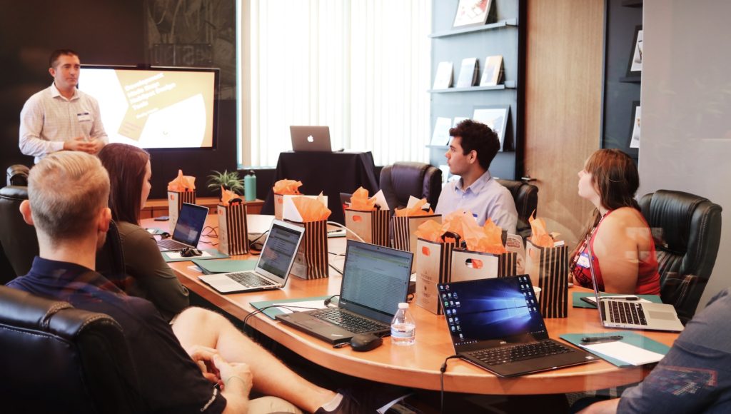 Uczestnicy spotkania siedzą przy jednym stole w biurze, przed nimi stoją laptopy.