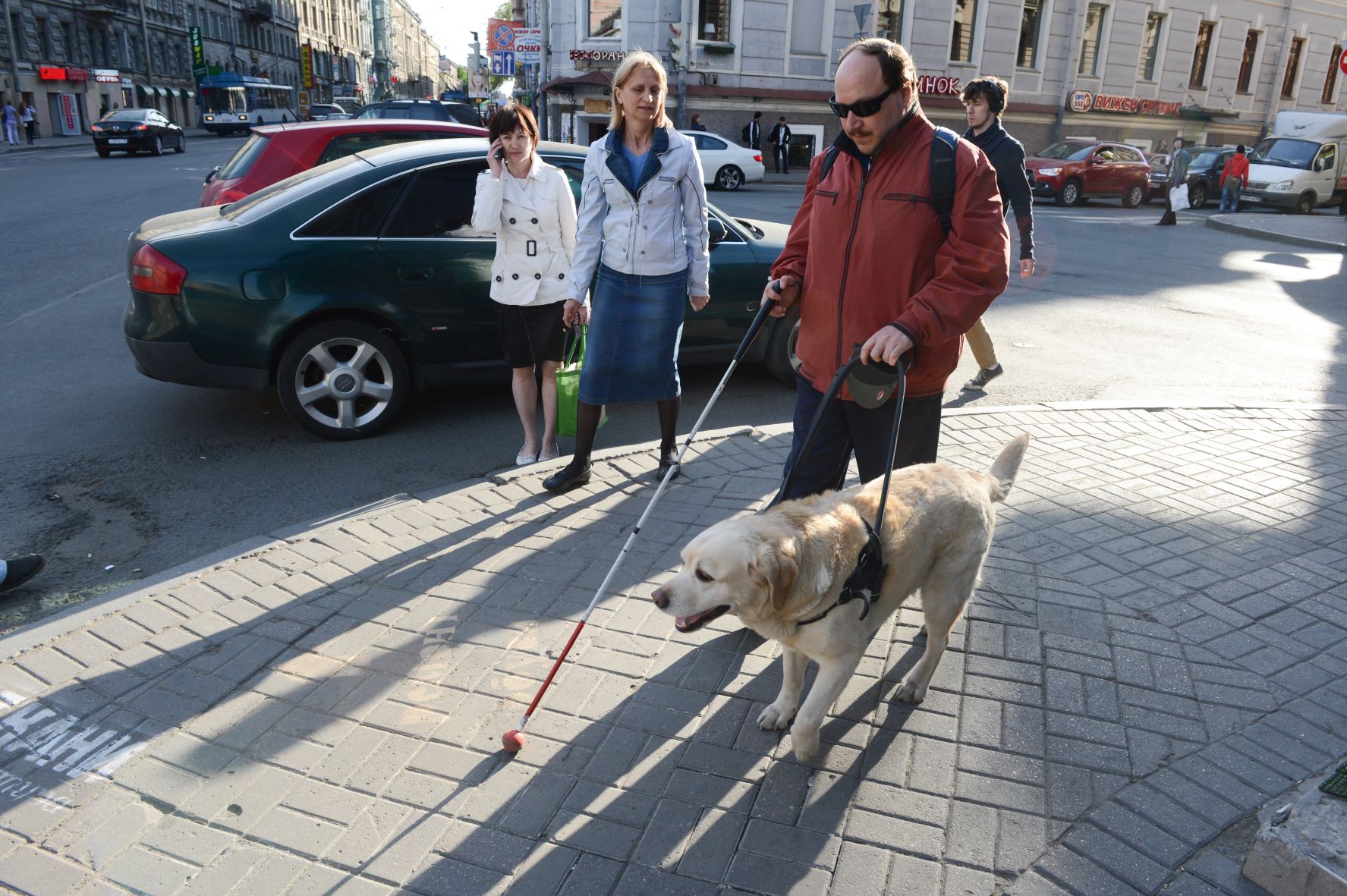 Niewidomy mężczyzna idzie po chodniku z psem przewodnikiem, obok przechodzą dwie kobiety