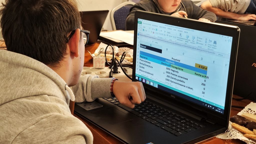 Młody mężczyzna siedzi przy laptopie i pracuje nad budżetem w pliku Excel. Zdjęcie zrobione zza jego pleców.