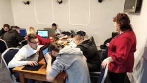 Uczestnicy projektu siedzą przy stole, pracują na laptopach. Wśród nich stoi prowadząca warsztaty.