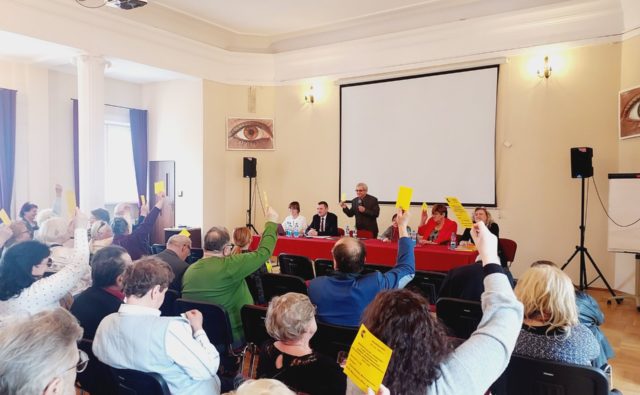 członkowie spotkania siedzą na sali i głosują; część z uczestników ma podniesione do góry żółte kartki