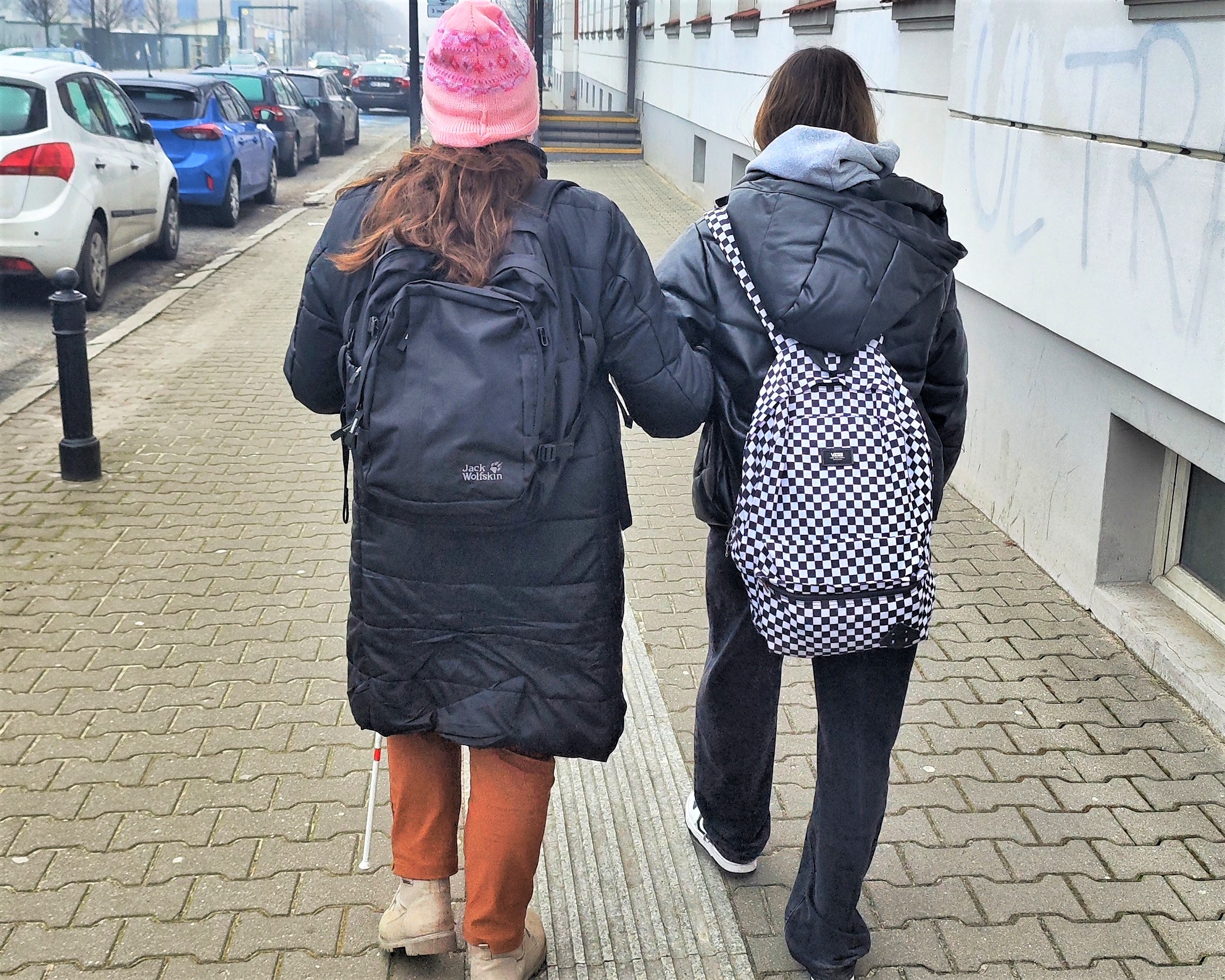 Kobieta niewidoma jest prowadzona przez młodą dziewczynę z plecakiem trasą po chodniku