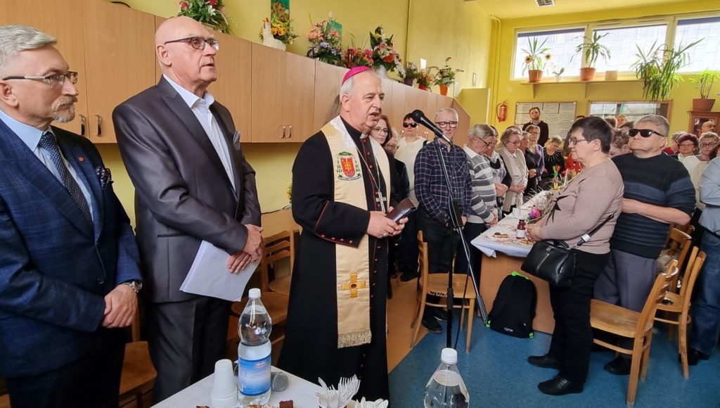 zaproszeni goście stoją przy stolikach, na pierwszym planie biskup mówi coś do mikrofonu, obok niego stoi prezes koła