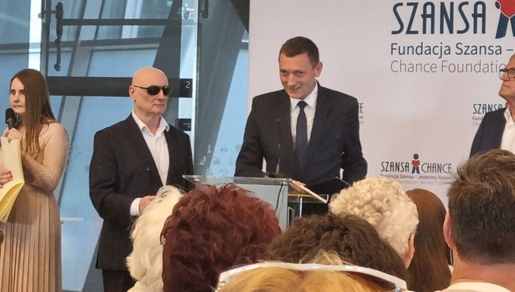 Andrzej Brzeziński stoi przed mównicą ze statuetką i przemawia do uczestników; po jego lewej stronie stoi Marek Kalbarczyk