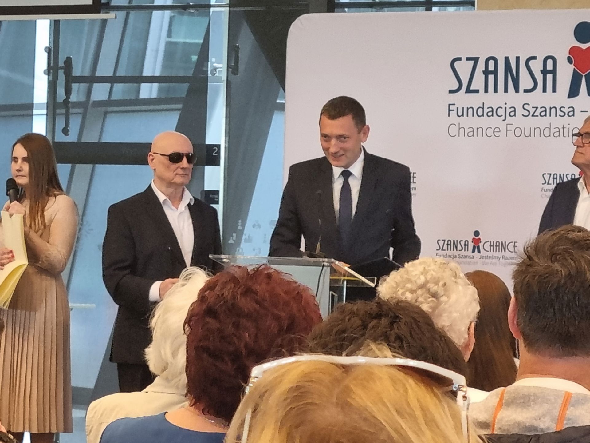 Andrzej Brzeziński stoi przed mównicą ze statuetką i przemawia do uczestników; po jego lewej stronie stoi Marek Kalbarczyk
