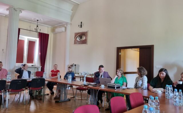 widok na salę, w której odbywa się spotkanie; przy stołach siedzą uczestnicy, prezes Andrzej Brzeziński mówi przez mikrofon i otwiera spotkanie