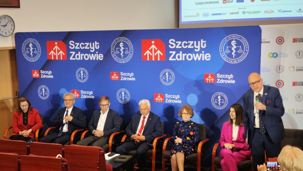 prelegenci siedzą przed uczestnikami, po prawej stoi moderator dyskusji; za nimi baner z nazwą wydarzenia: "Szczyt Zdrowie"