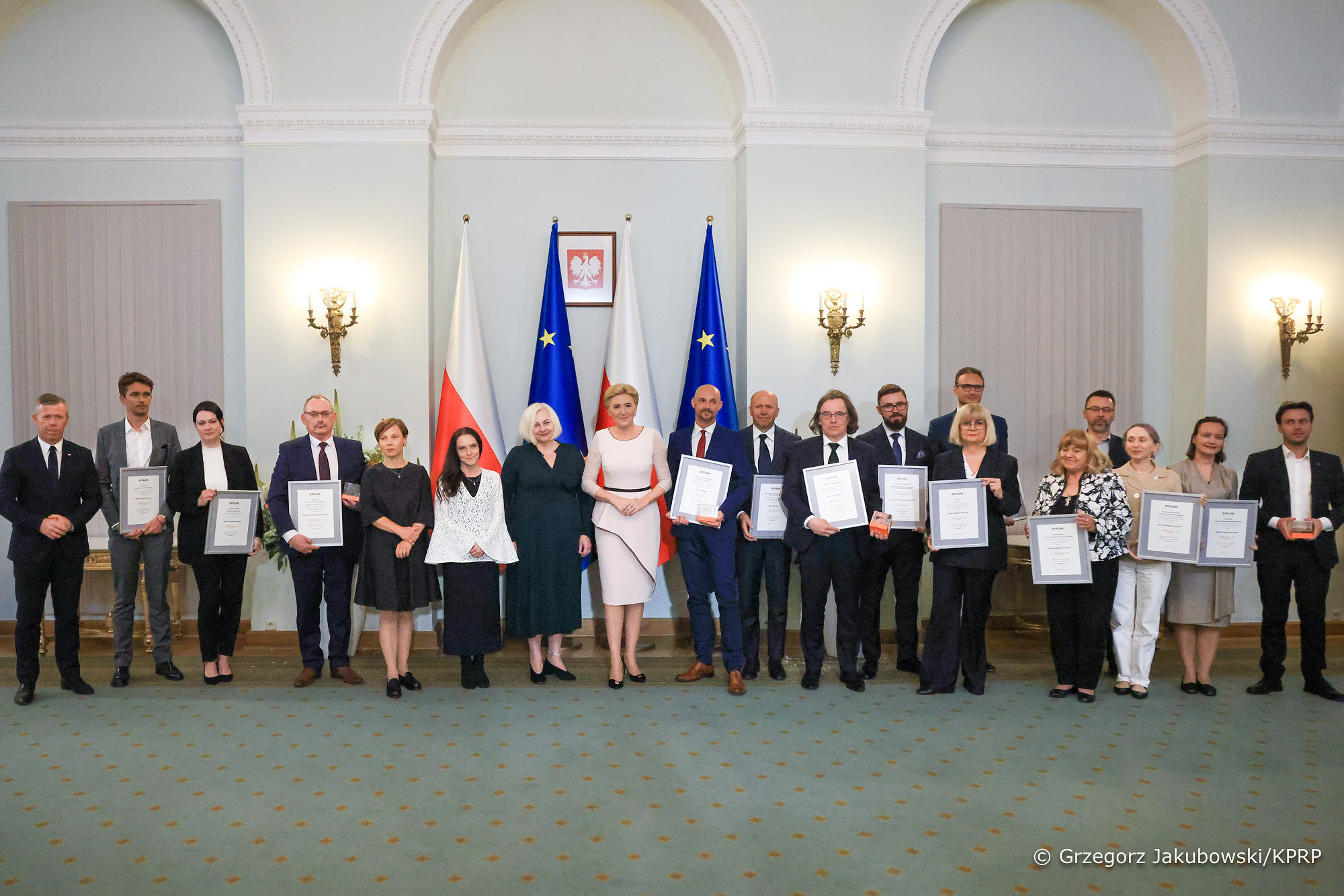 Pierwsza Dama, Agata Kornhauser-Duda stoi w Pałacu Prezydenckim, wokół niej stoją laureaci konkursu z dyplomami i statuetkami.